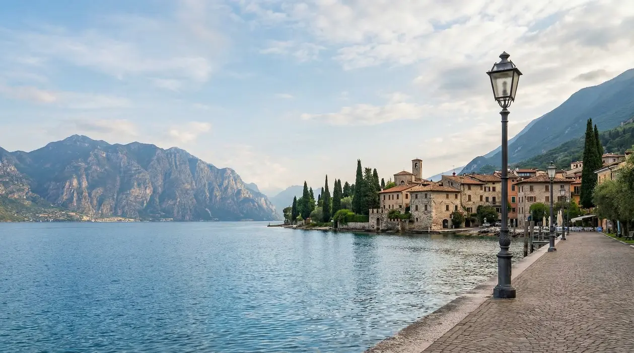 Lungolago del Lago di Garda con borgo storico, cipressi e montagne sullo sfondo in una giornata tranquilla